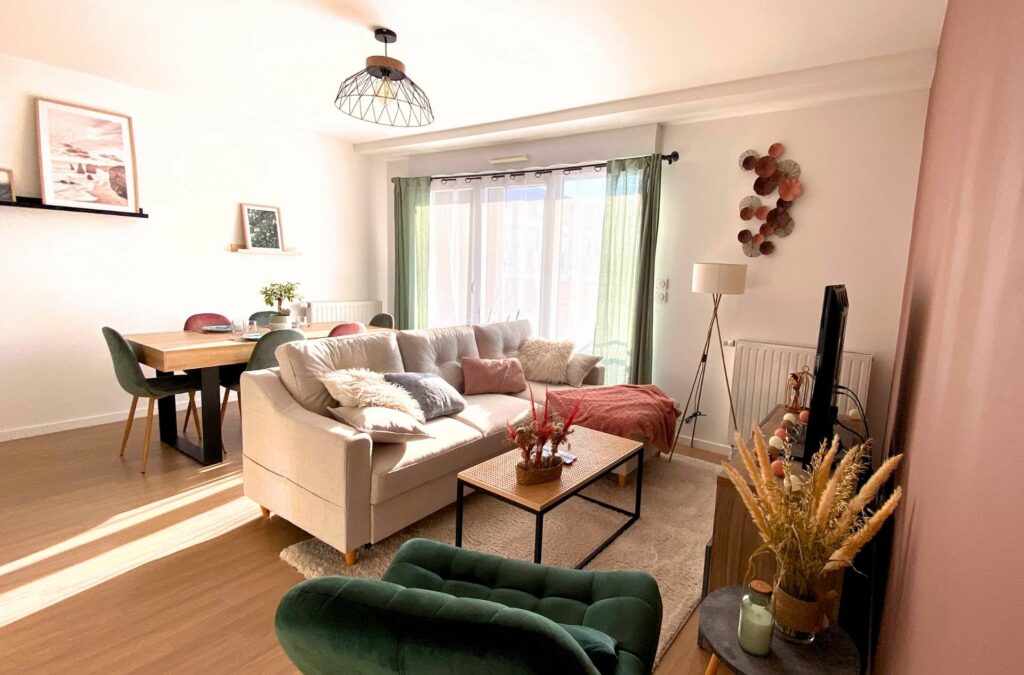 Un séjour lumineux avec une ambiance pleine de douceur, un canapé beige en velours, un fauteuil en velours vert sapin, des coussins rose et un mur peint en couleur vieux rose