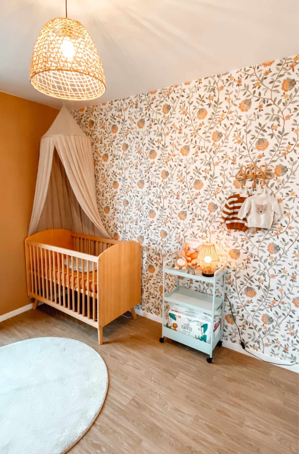 Une chambre pour bébé garçon ou fille, à l'ambiance naturelle et douce. Le mobilier est en bois et en métal peint en vert d'eau. Un tapis moelleux et un papier peint floral. 